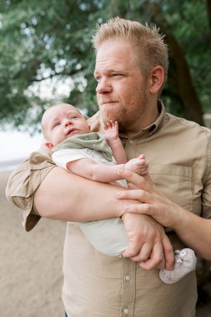 A tender moment captured as adult figure holds tiny yawning baby wearing light colored clothing outdoors.