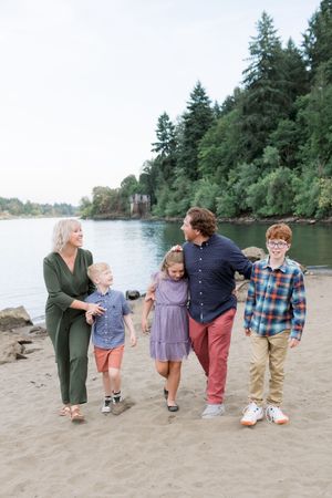 Family enjoying a casual walk along a scenic riverbank with evergreen trees in the background on a cloudy day.