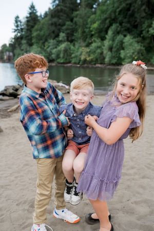 Children playing and laughing together on a sandy beach with evergreen trees and water in the background.