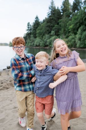Three children laughing and playing together on a beach with evergreen trees in the background at dusk.