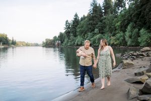People walk and play along the water's edge of a scenic river with a wooded shoreline.