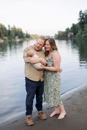 A couple embraces while holding a newborn baby on a peaceful lakeshore with trees in the background.