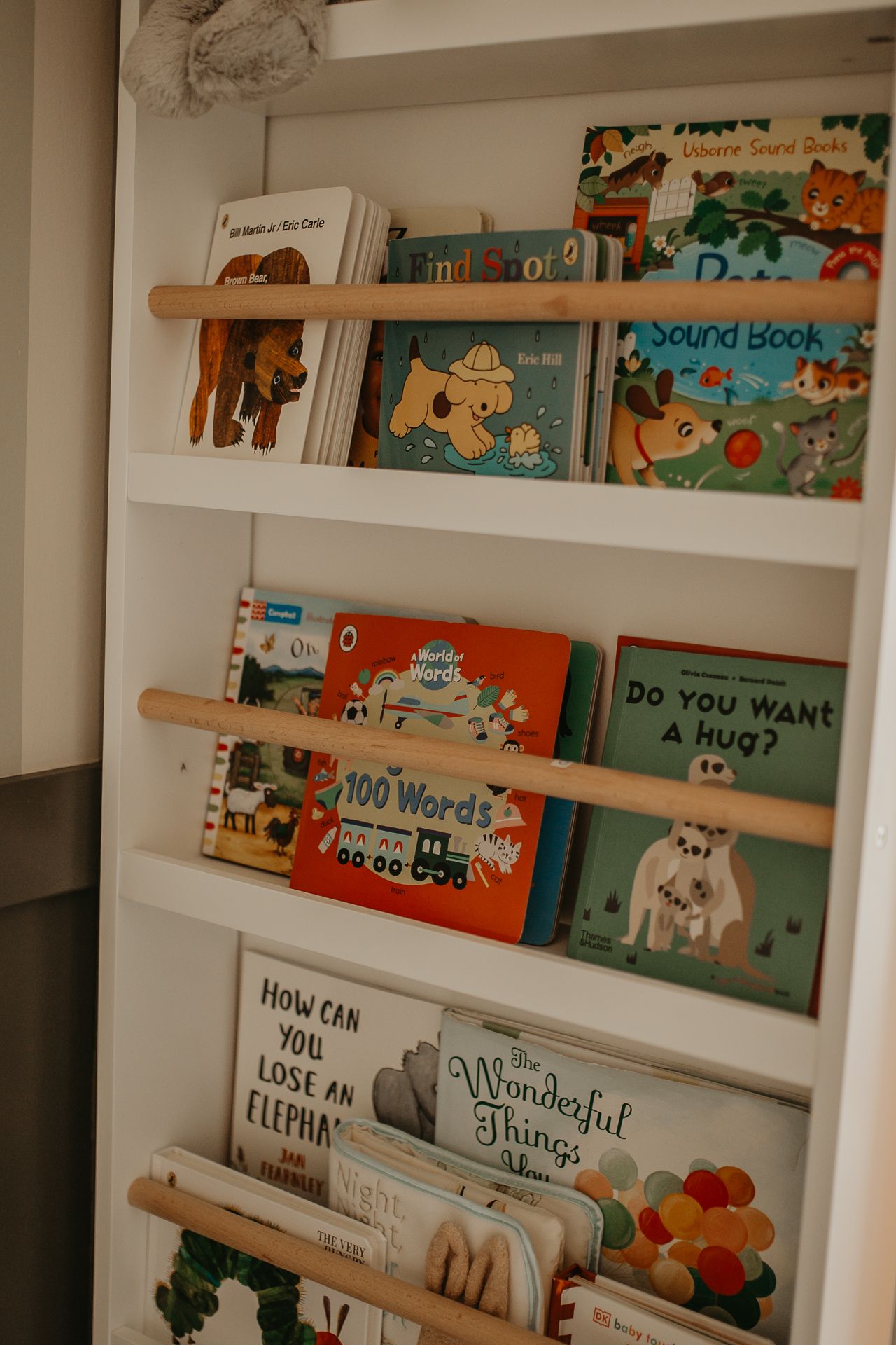 White wooden floating shelves display a collection of children's books with colorful spines against a light colored wall.