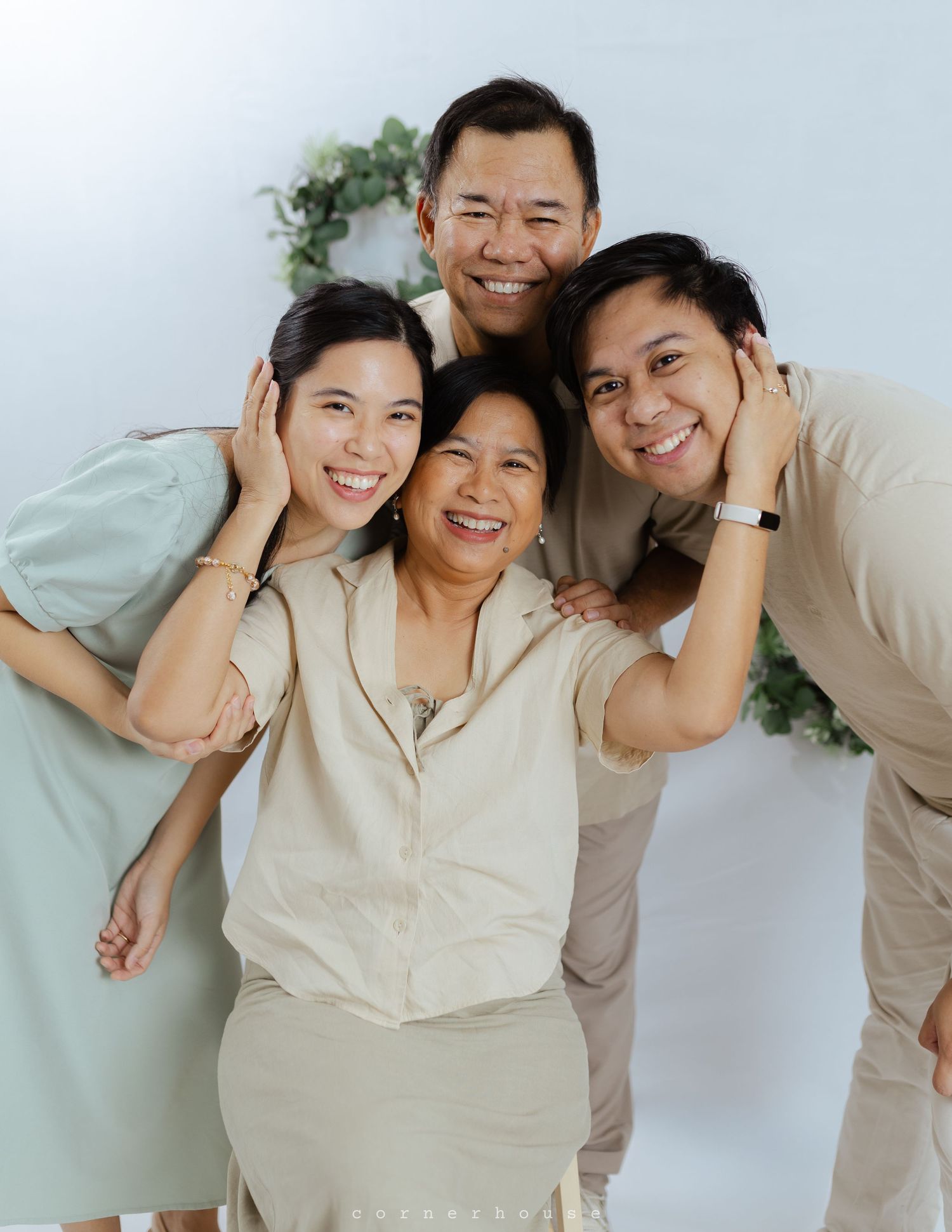 A family wearing neutral colored clothing poses together against a light background with greenery decoration.