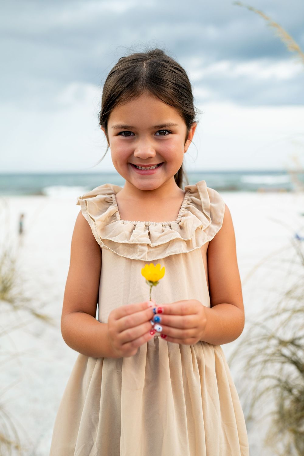 A child in a beige ruffled dress holds a yellow flower while standing on a beach with cloudy skies.