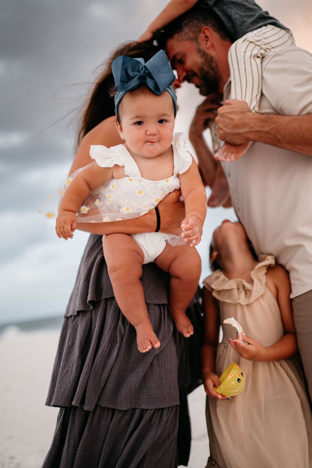Family enjoying tender moments together on a beach with a baby in white dress.