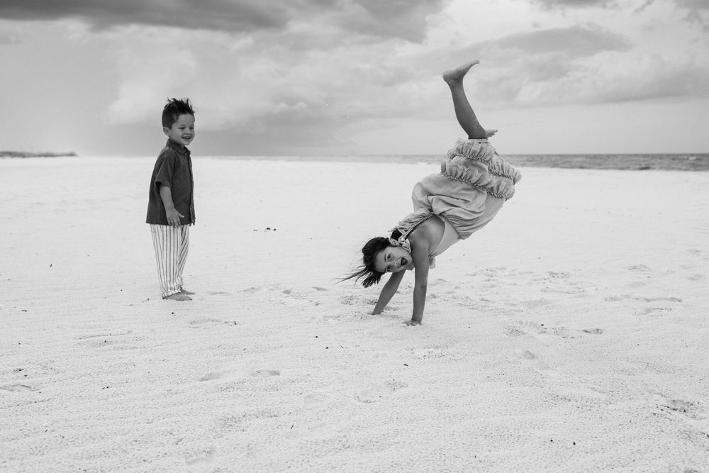 Two people playing on a white sandy beach with one doing a handstand while another watches under cloudy skies.