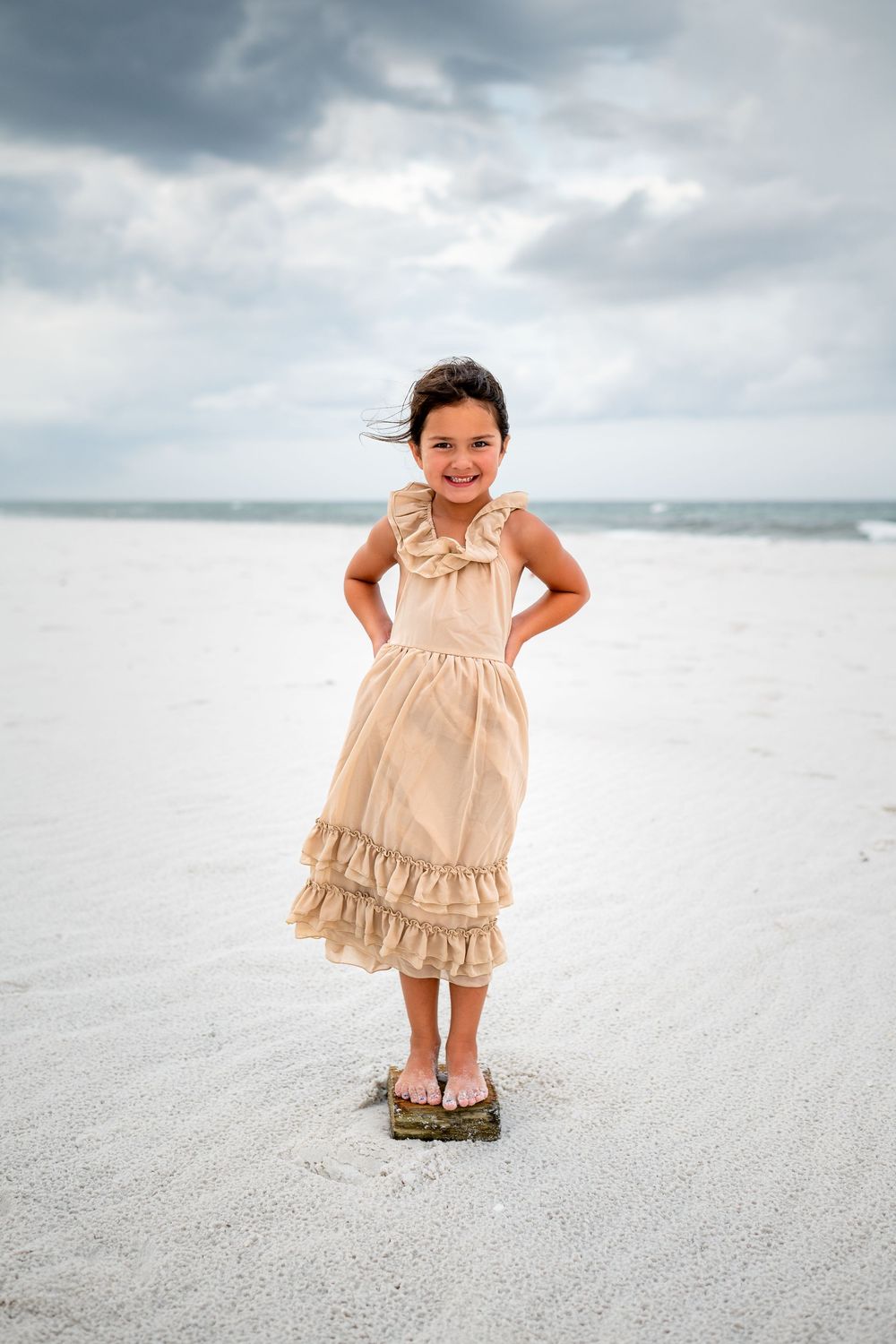 A child wearing a ruffled cream-colored dress stands on pristine white sand at the beach.