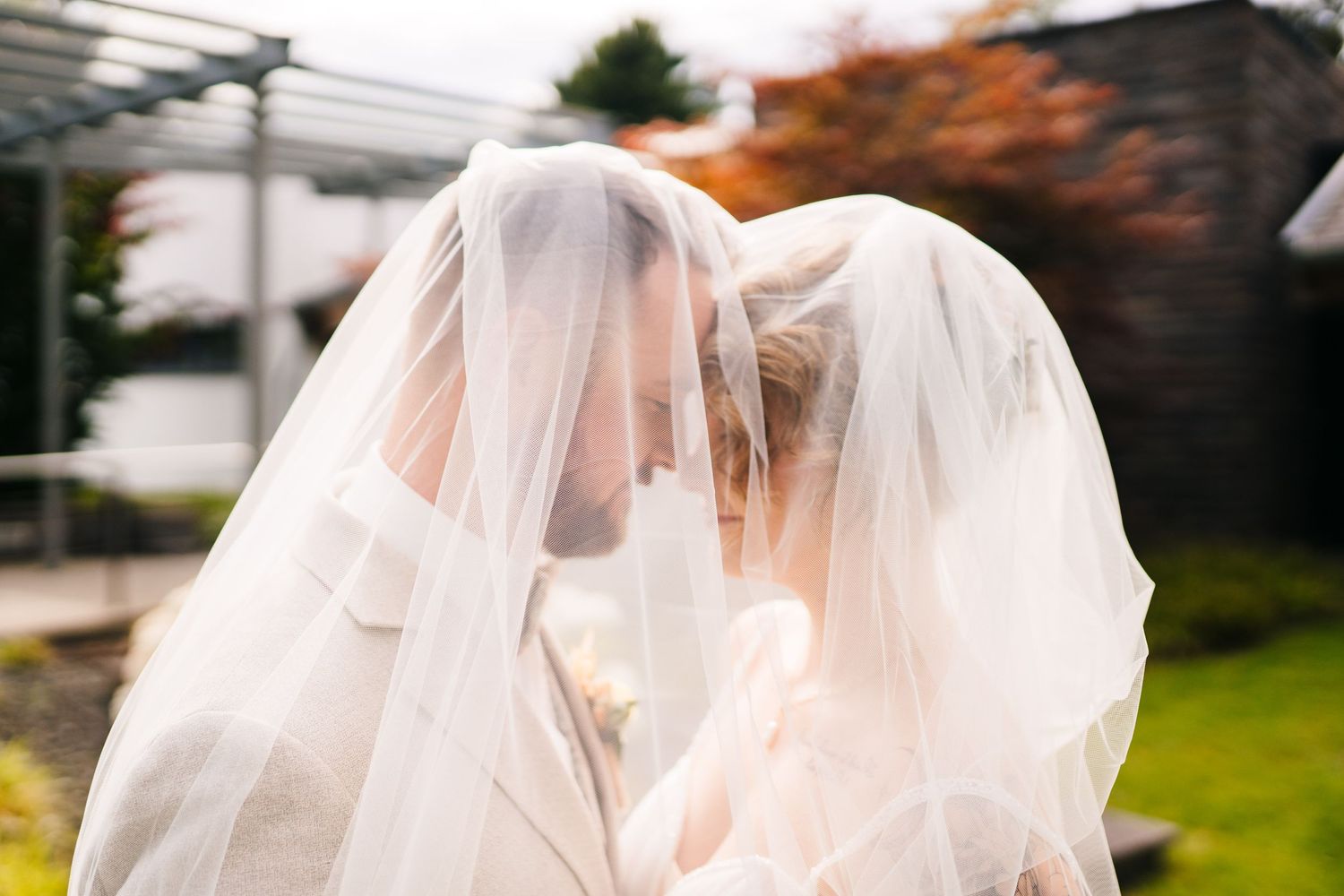 Two figures share a tender moment under flowing white veils in a garden setting.