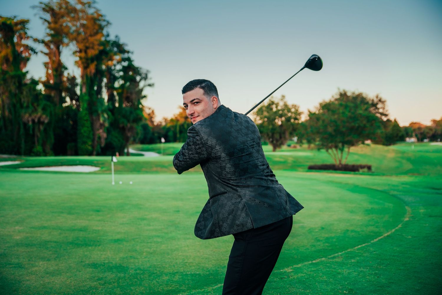 Man in dark suit swinging golf club on lush green golf course at sunset.