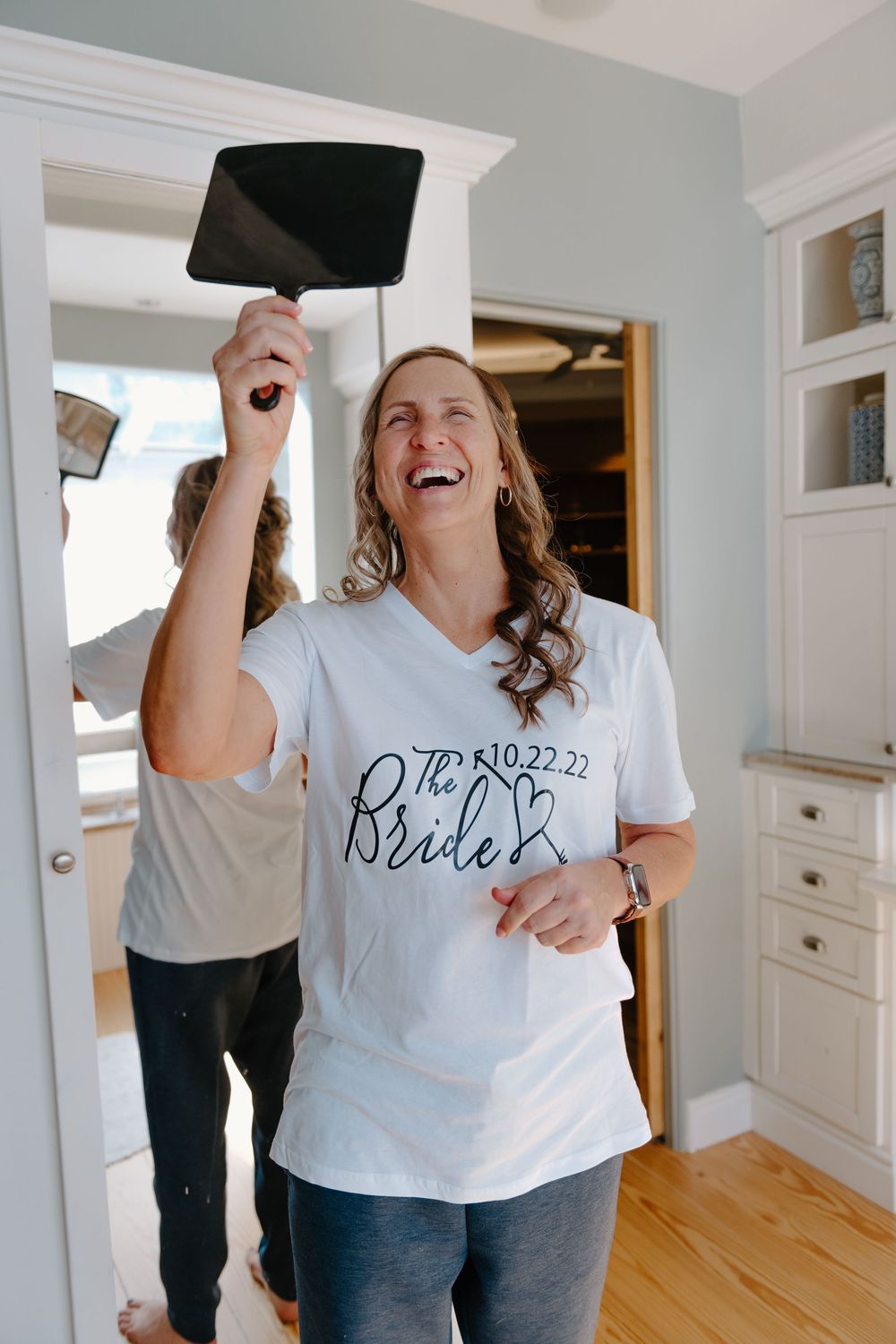 A person in a white t-shirt with text design laughs joyfully while holding up a graduation cap in an indoor setting.