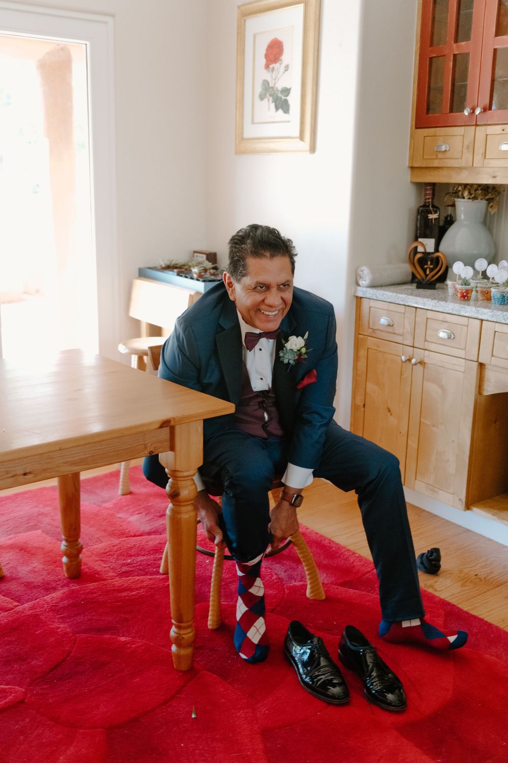 Someone in a navy suit sits on a wooden table with colorful socks and dress shoes on a red carpet.
