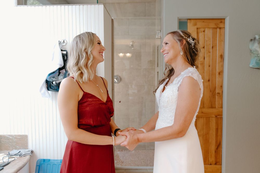 A bride in a white gown and companion in burgundy dress share a joyful moment while getting ready.
