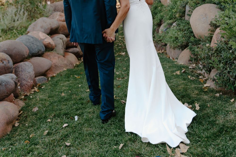 A couple in wedding attire stands together on grass near rustic logs during an outdoor photoshoot.