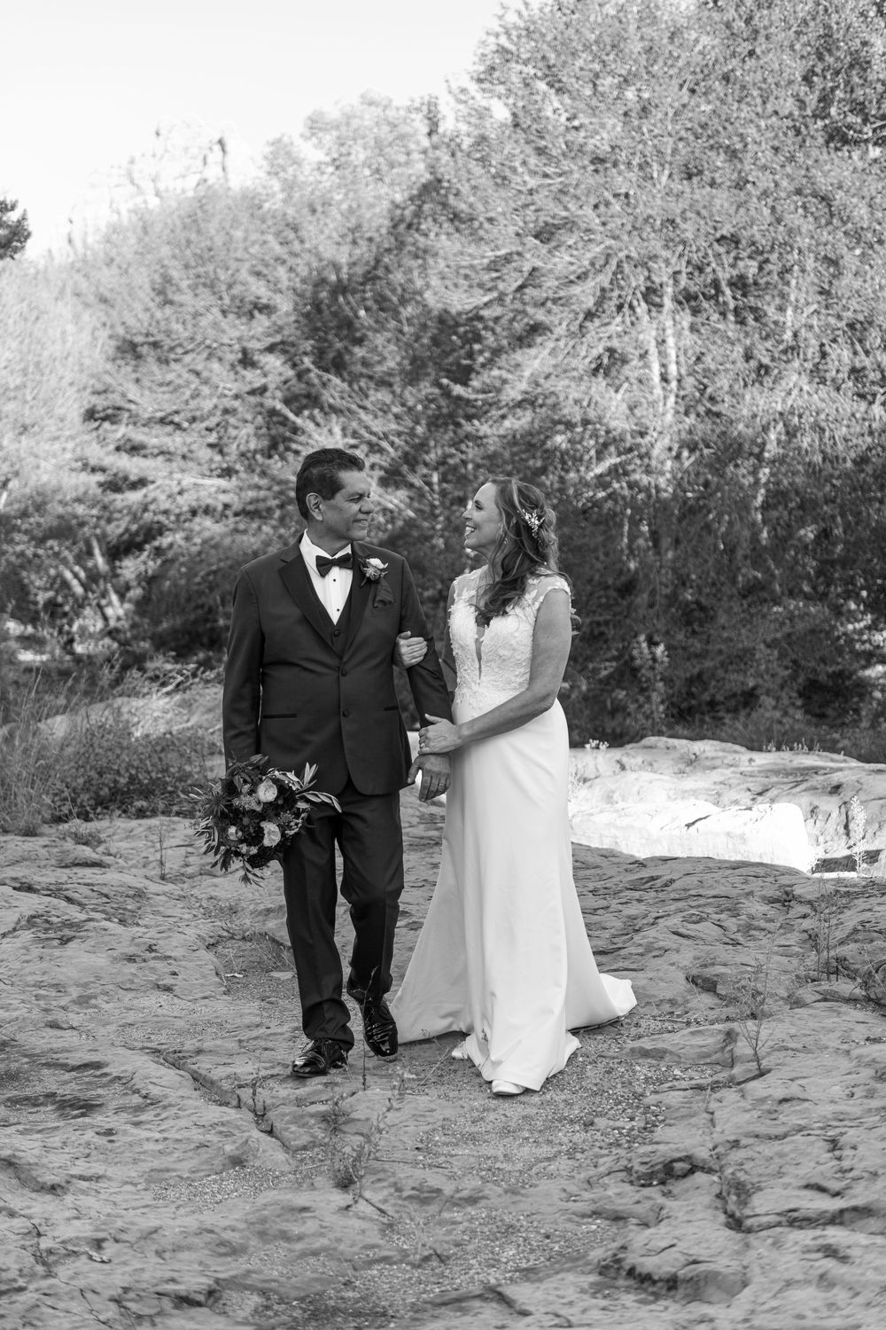 A couple in formal wedding attire walks along a rocky beach path in black and white.