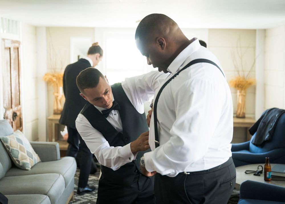 Two people help adjust formalwear and accessories while getting ready in a bright hotel room.