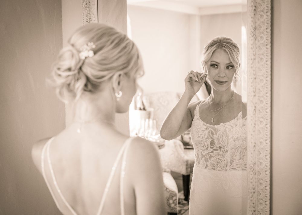 A reflection of someone in a white dress looking in a mirror while putting on earrings.