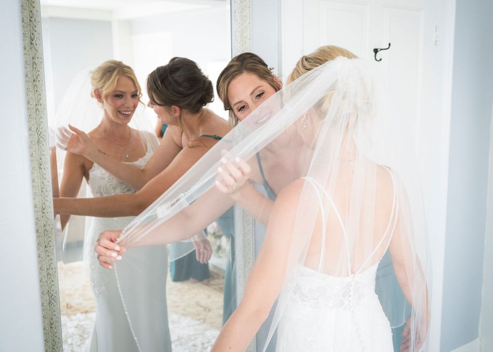 Wedding party members help adjust a bride's veil in a bright room.