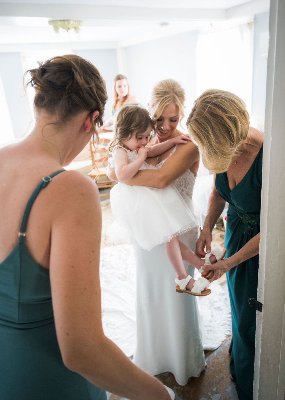 Wedding party helps bride with final dress adjustments in sunlit room with white curtains.