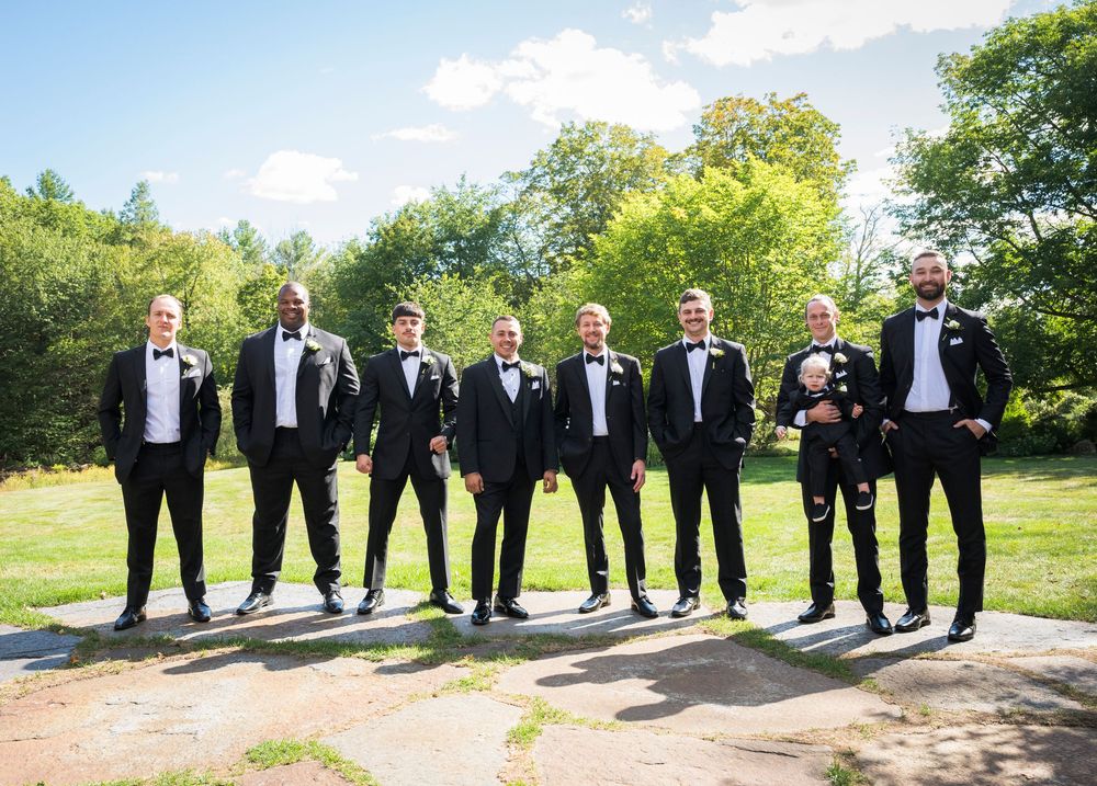 Groomsmen in formal black tuxedos lined up outdoors for wedding party photos.