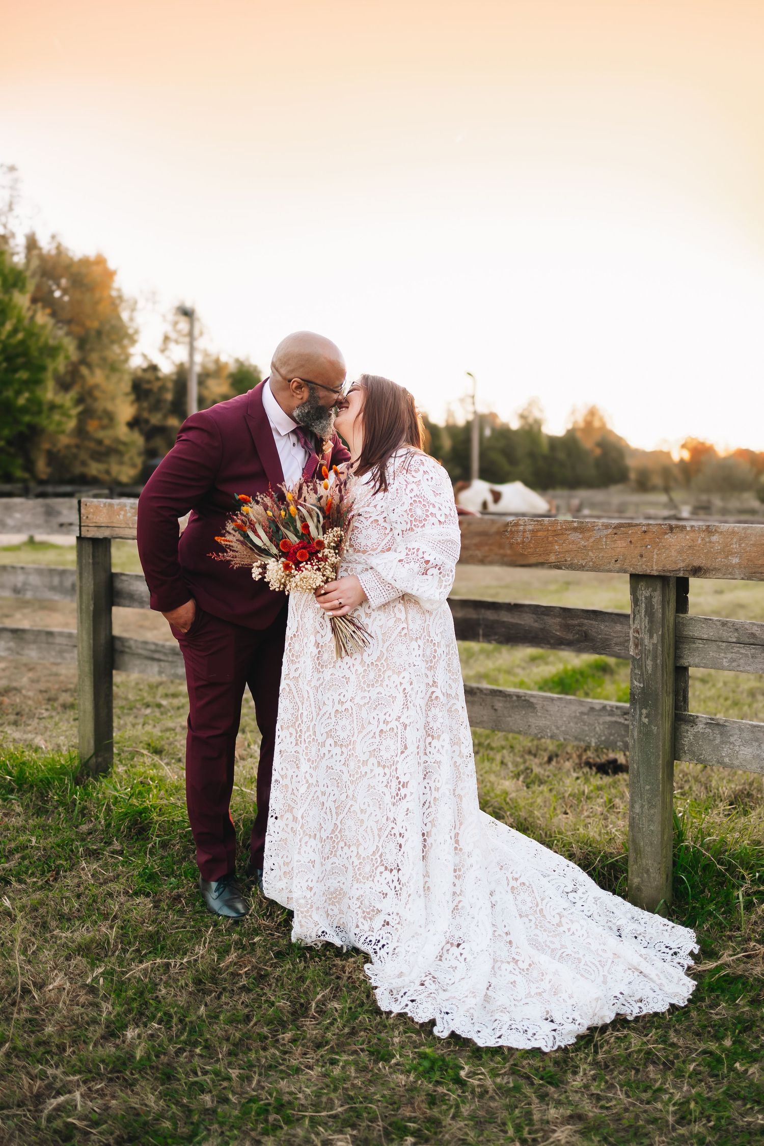 A romantic wedding couple kisses at sunset against a rustic wooden fence.