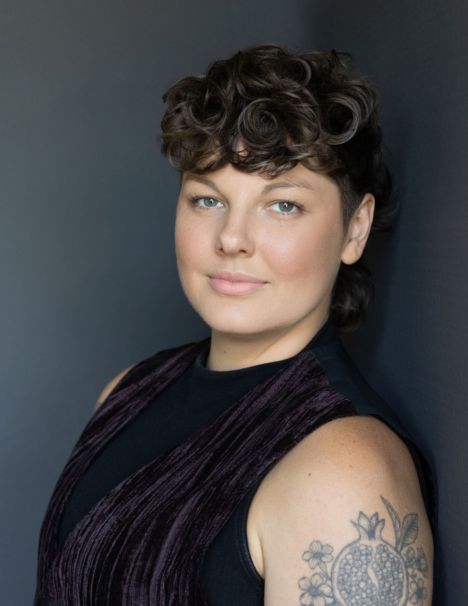 Close-up headshot of Theo Hayes, a non-binary opera singer, against grey backdrop, wearing a dark collared shirt, short styled hair.