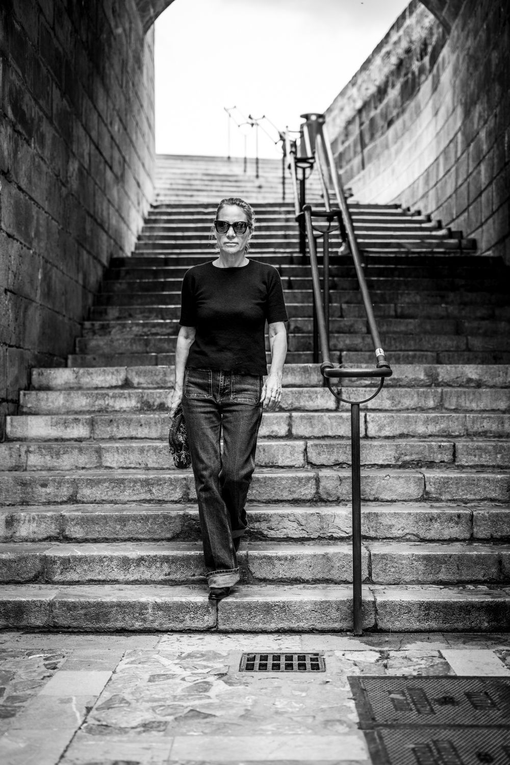 Schwarz-Weiß-Modefoto einer Frau mit Jeans, schwarzem Pullover und Sonnenbrille auf Treppen in Palma