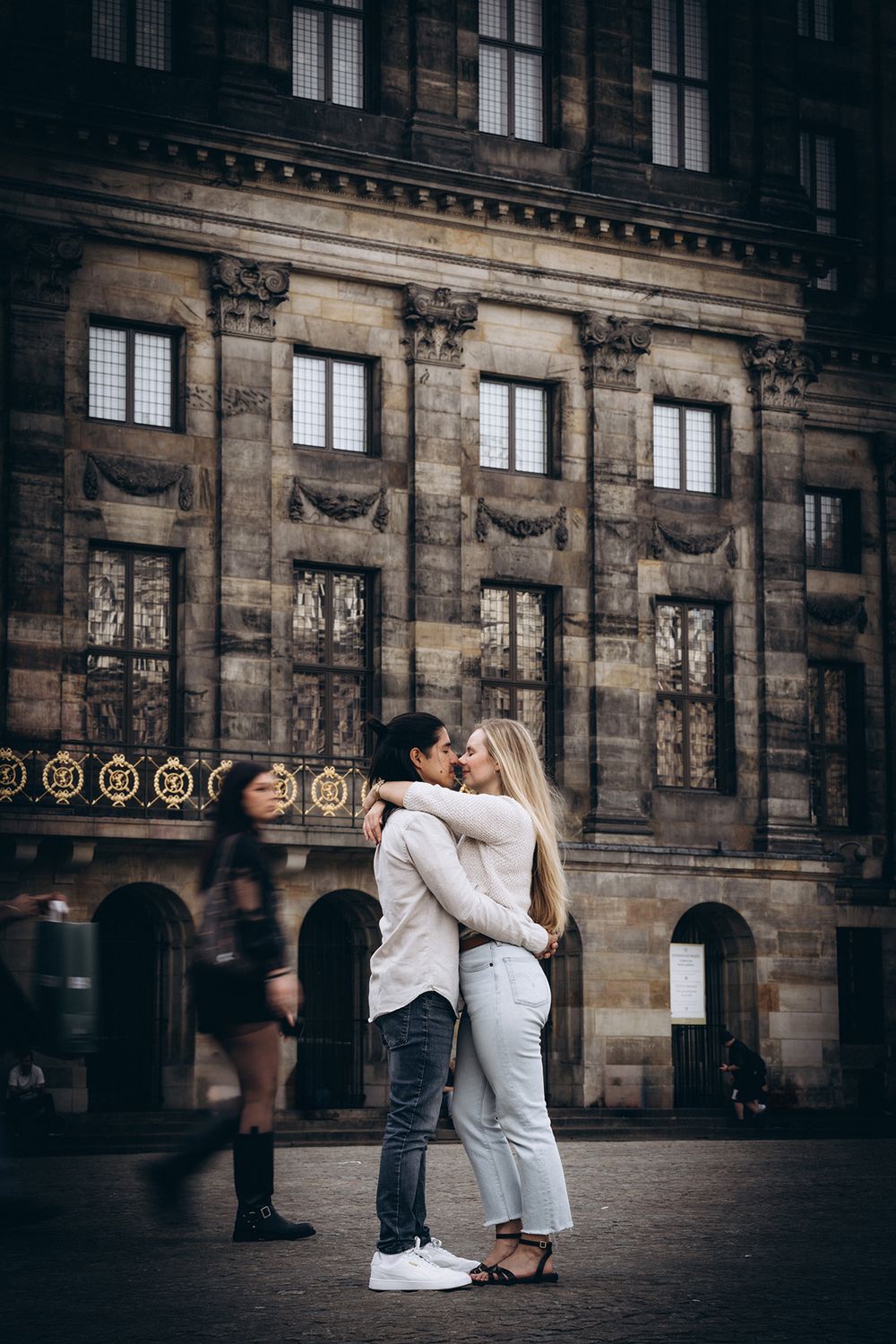 Paar steht ruhig in Amsterdam, während Passanten unscharf vorbeigehen. Urbane Paarfotografie im Hochformat.