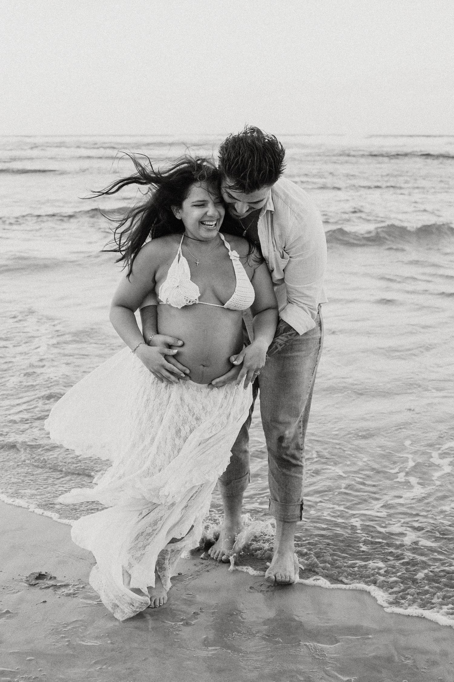 A couple embraces while standing in the ocean waves, wearing casual beach attire.