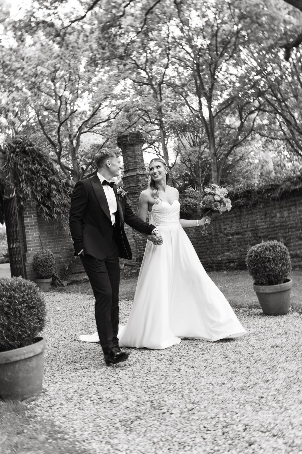 A black and white wedding photo showing a couple outdoors in a garden setting with brick walls and potted plants.