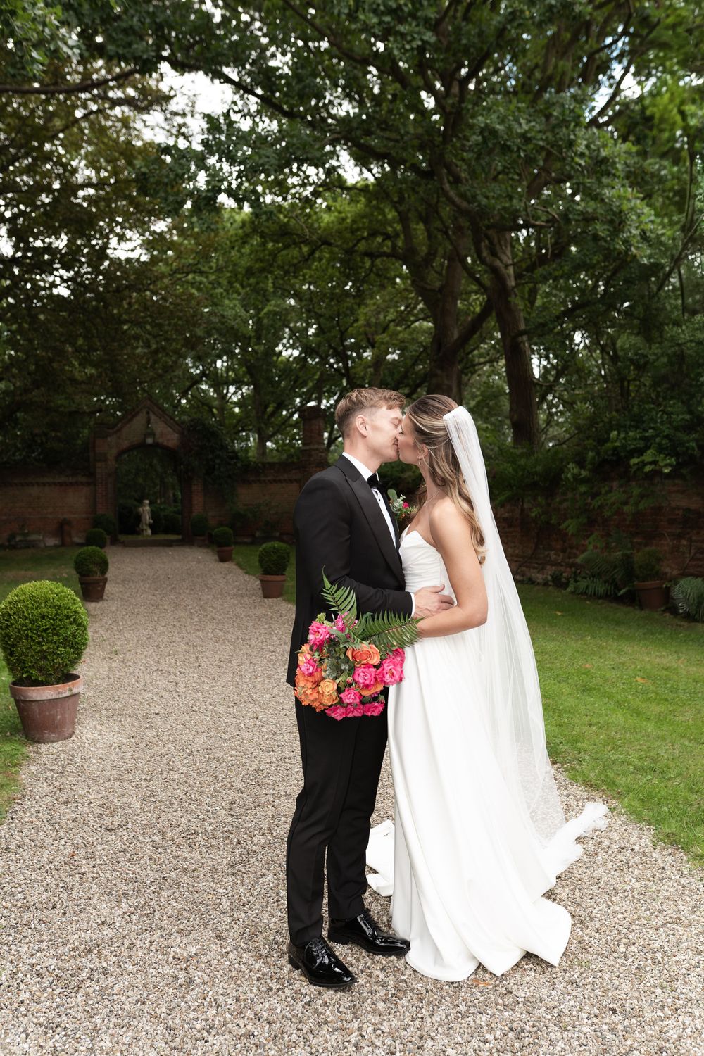 A couple shares an intimate moment on a gravel path surrounded by lush greenery and flowering plants.