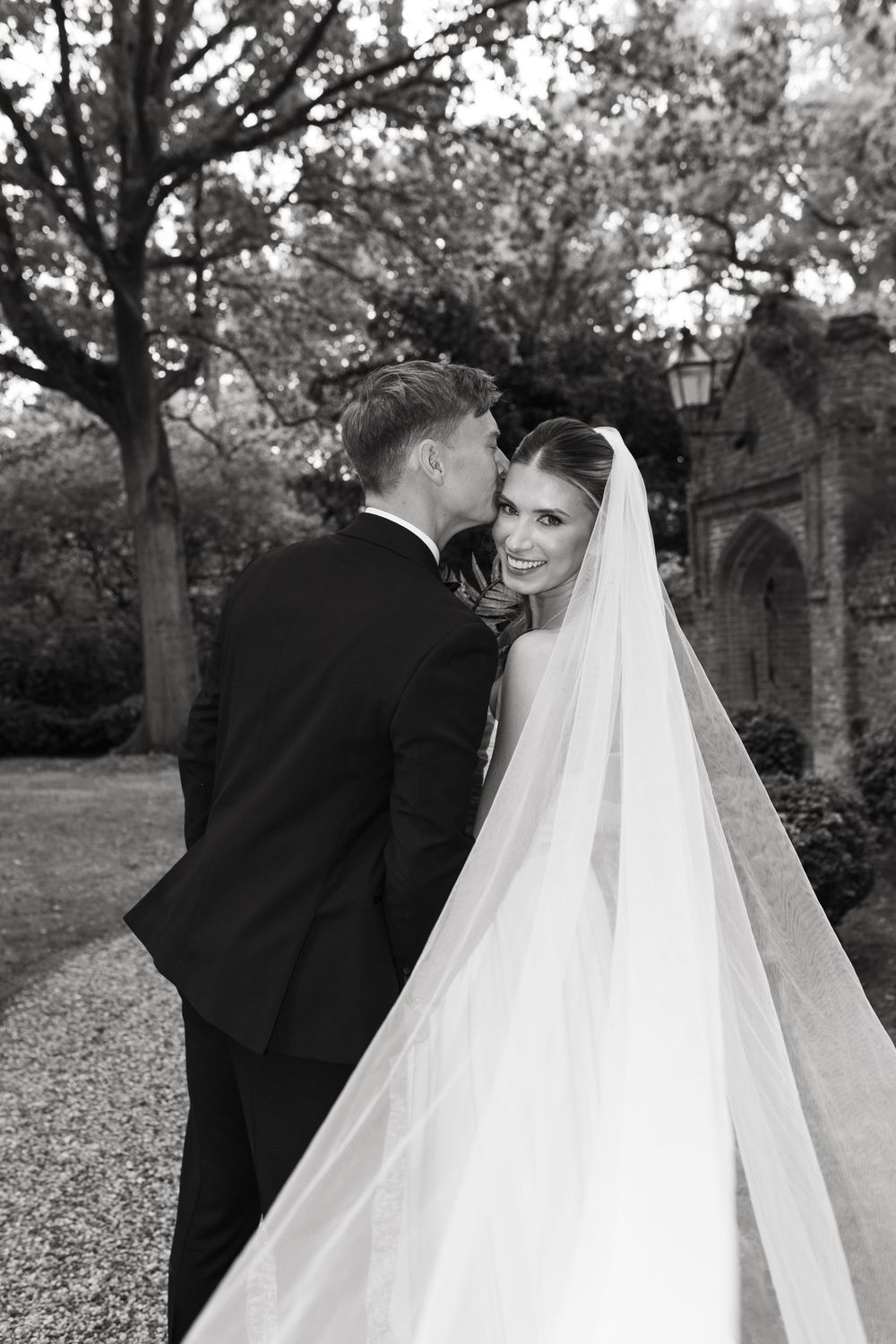 A wedding couple shares a tender moment outdoors with a flowing white veil against a backdrop of trees.