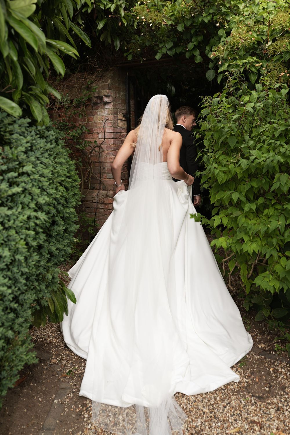 A bride in a flowing white wedding dress and veil walks along a garden path beside lush green foliage and brick walls.