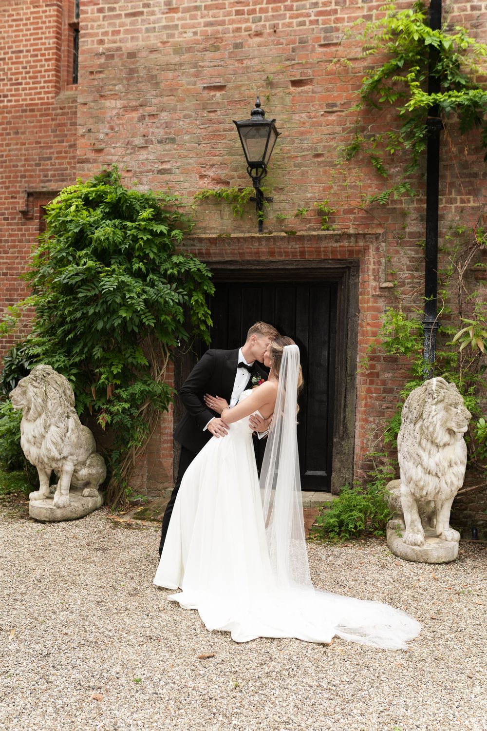 A romantic wedding photoshoot in front of a brick wall with stone lion statues and gravel path.