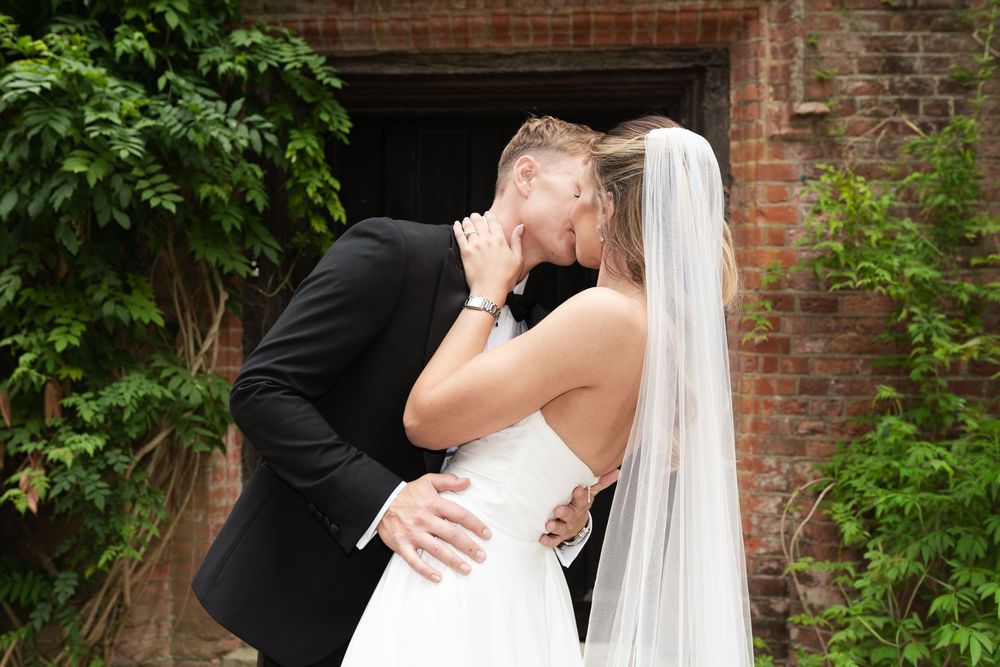 A bride and groom share romantic kisses in a lush garden setting with brick walls.