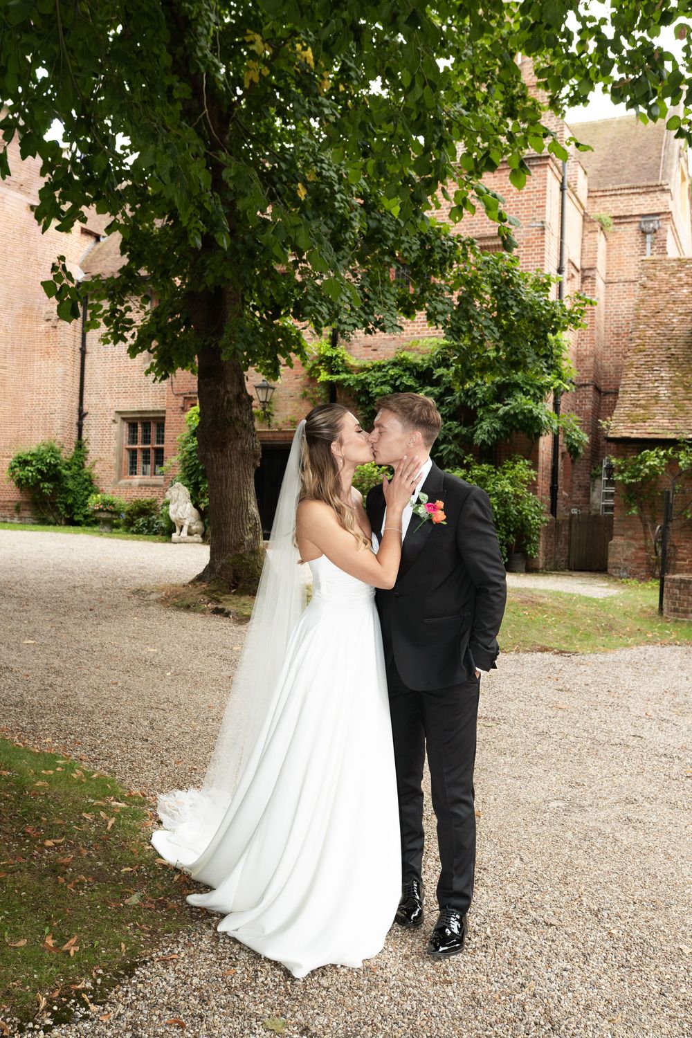 Bride and groom walking along a tree-lined gravel path beside historic brick buildings.