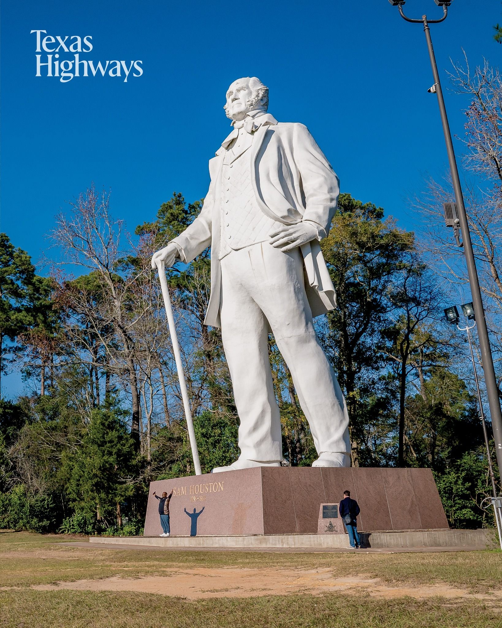 Giant white statue known as Sam Houston stands tall against blue sky in Huntsville, Texas with trees in background.