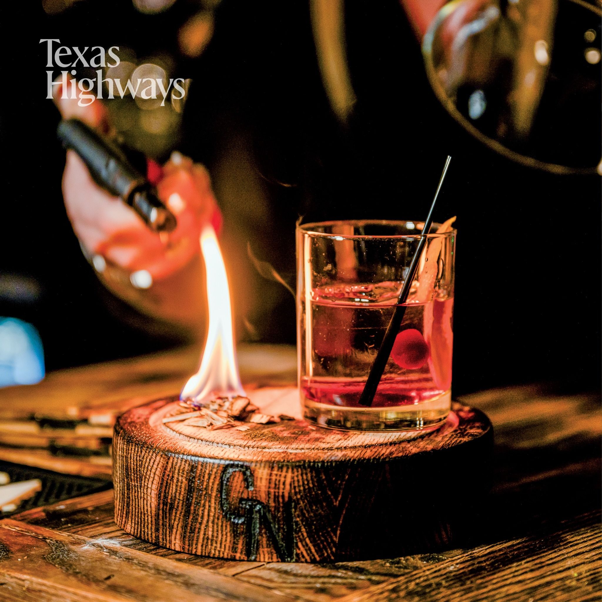 A cocktail served in a rocks glass sits on a rustic wood slice with a flame burning beside it in dim lighting.