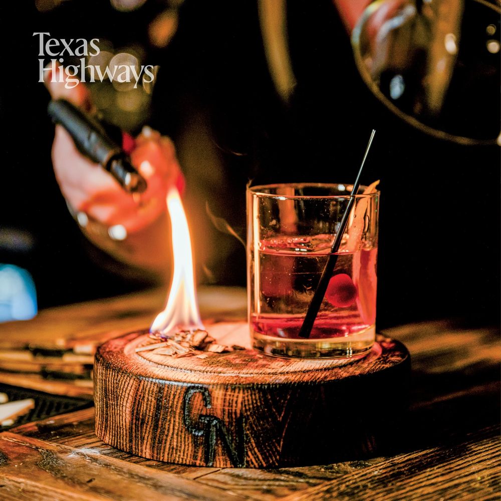 A cocktail served in a rocks glass sits on a rustic wood slice with a flame burning beside it in dim lighting.