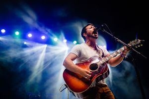 A performer plays acoustic guitar on stage with colorful blue lighting and fog effects in the background.