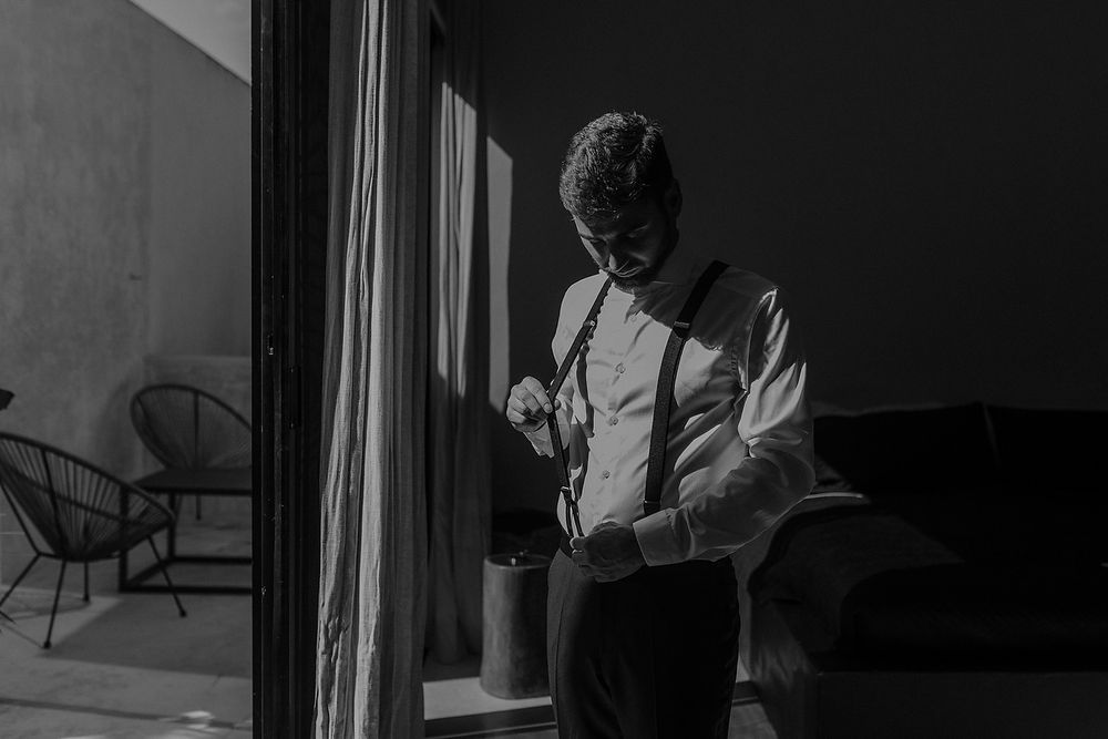 Black and white photo of a groom in suspenders and dress shirt preparing for his wedding day.