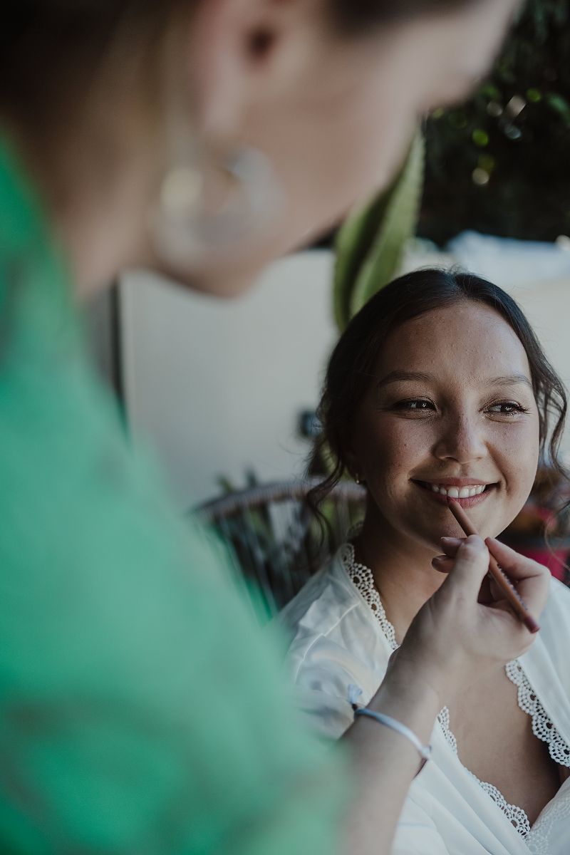 Bride smiling joyfully as makeup artist applies lipstick during wedding preparation in white lace dress.