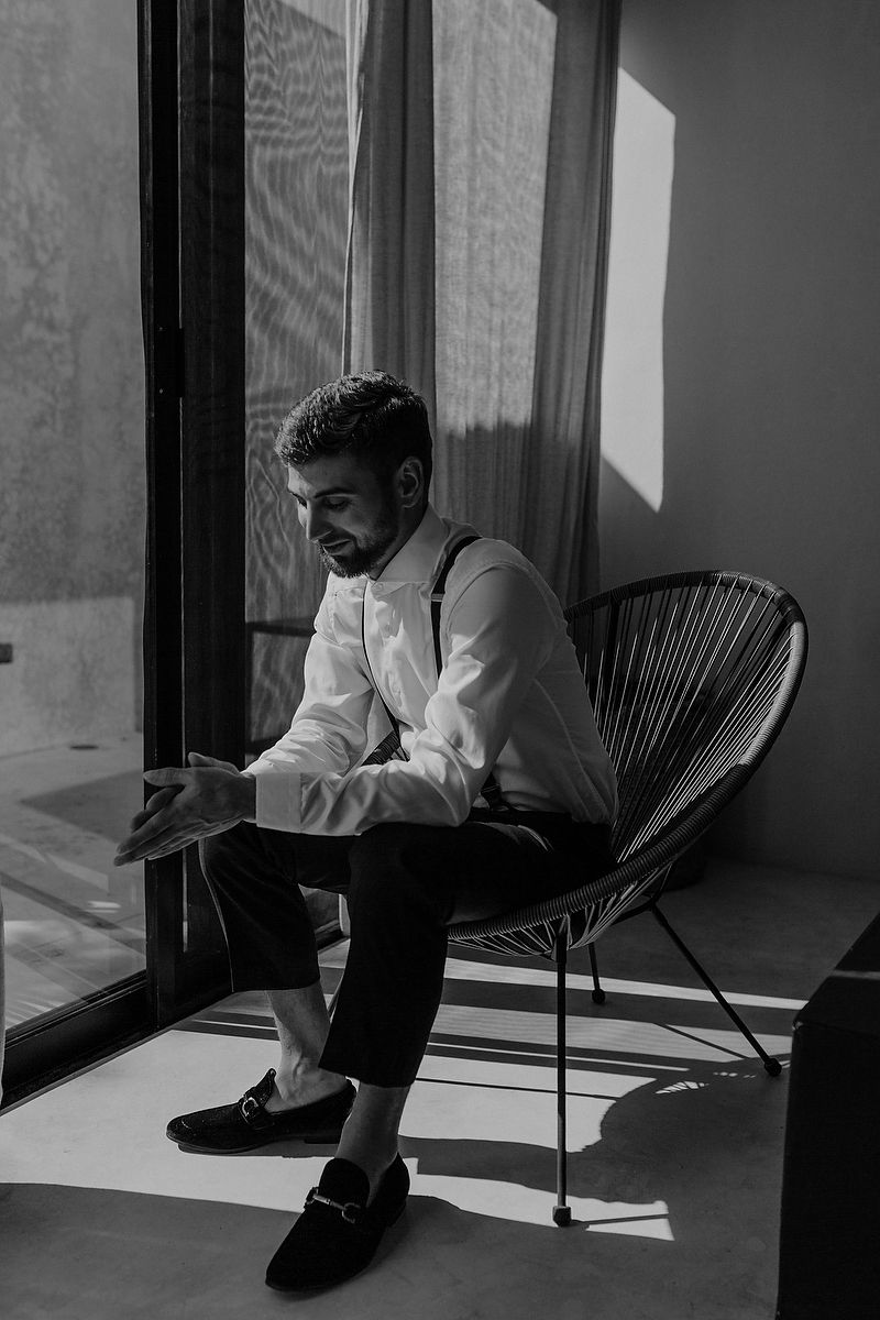 Black and white photo of a well-dressed man in suspenders sitting in a wicker chair by a sunlit window.