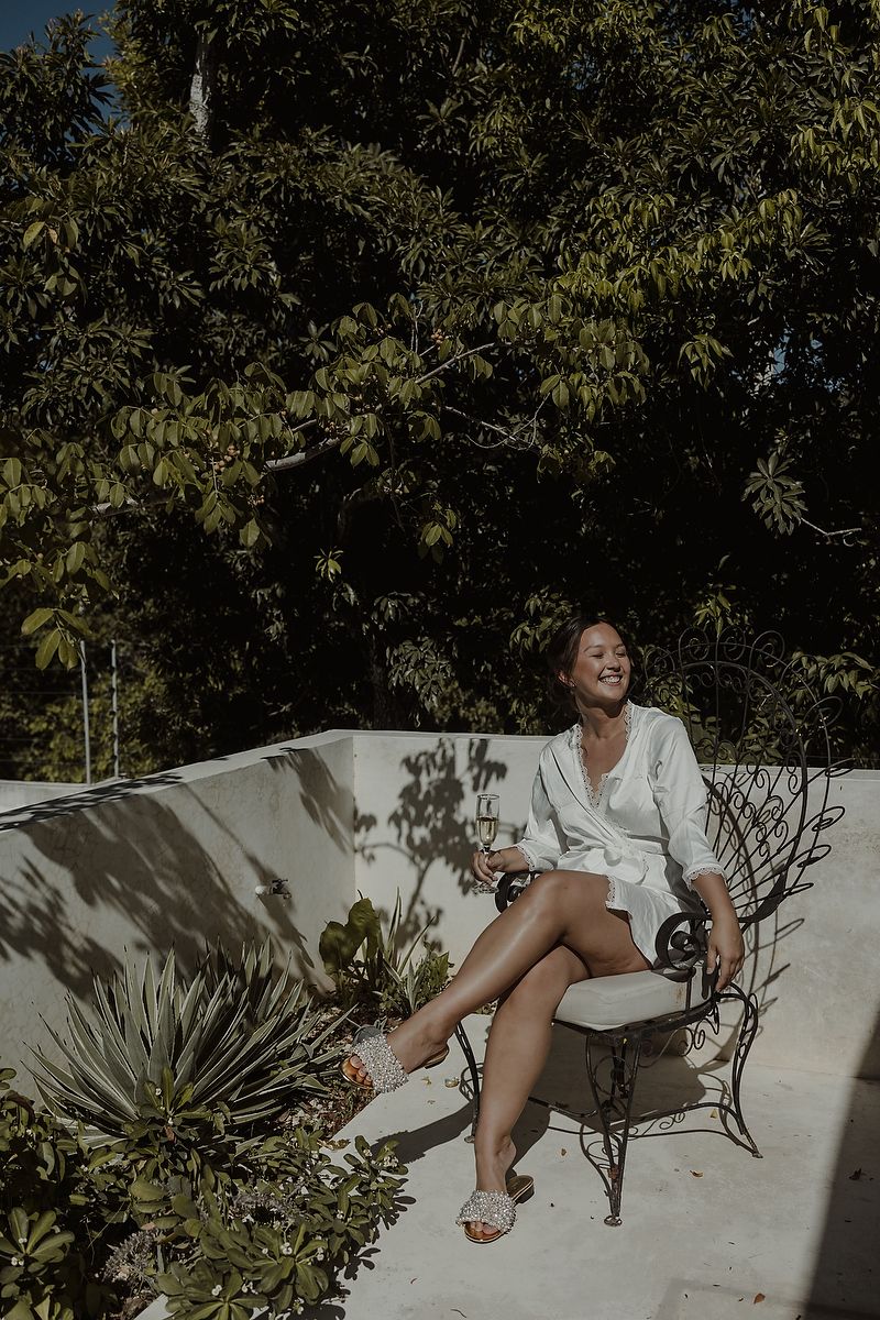 Woman in white dress sitting on ornate iron chair on sunny terrace surrounded by lush green tropical trees.