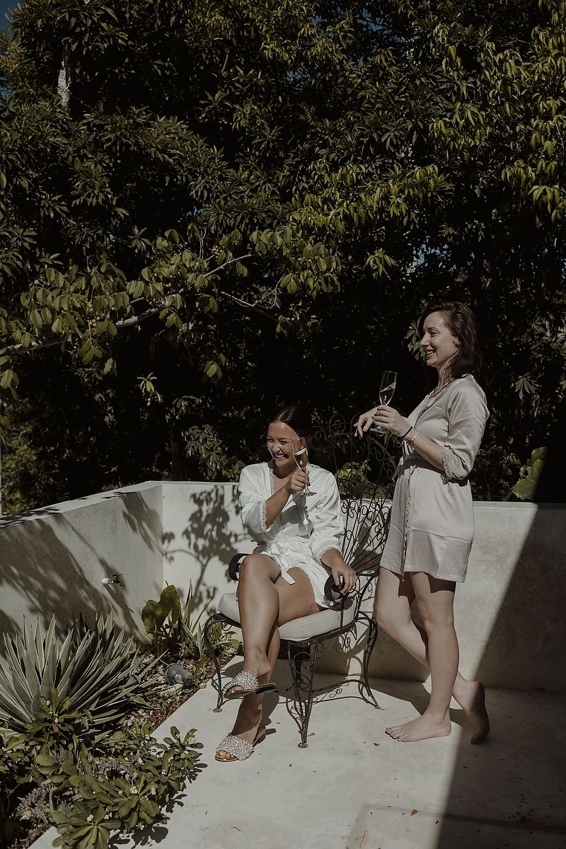 Two women in robes enjoy drinks on a sunny patio surrounded by lush green trees and tropical plants.