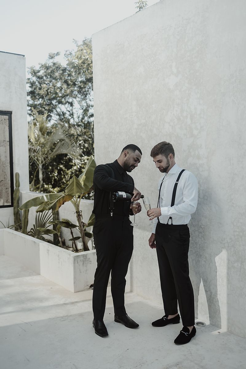 Two well-dressed men in black and white formal attire examine an item outdoors against a white wall.