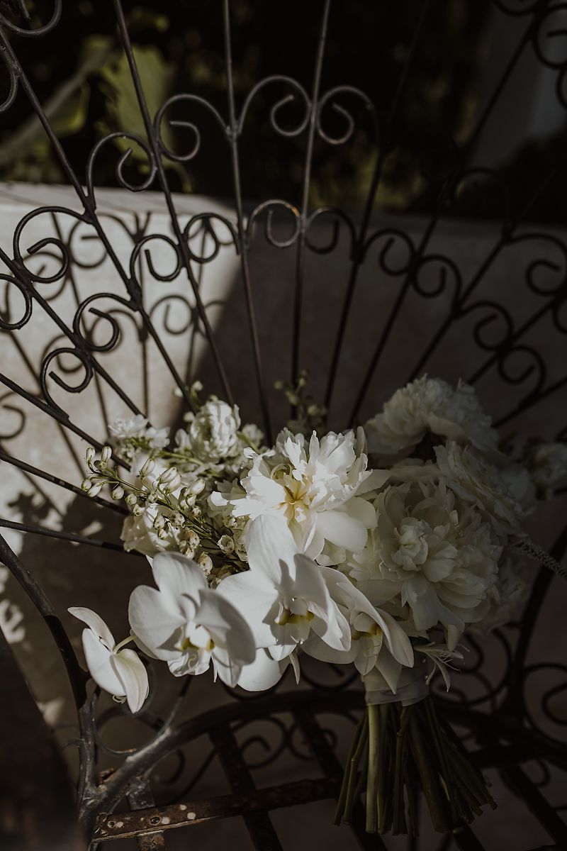 Elegant white wedding bouquet with peonies and orchids resting on ornate wrought iron chair.