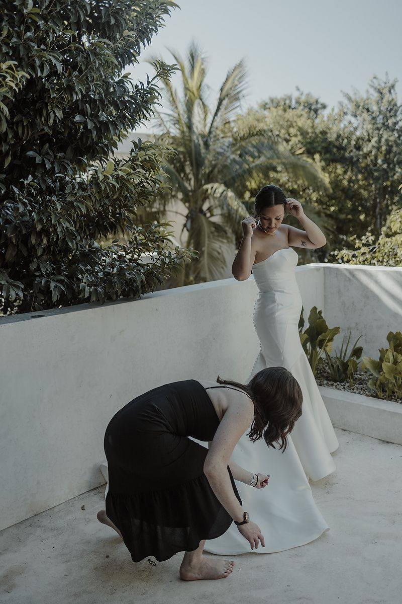 A bride in a white gown gets dress adjusted by assistant on a terrace surrounded by lush greenery.