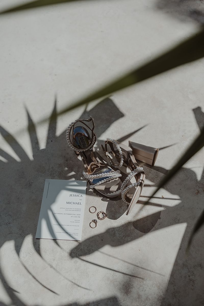 Wedding flat lay with invitation, rings, heels, and jewelry casting palm shadows on concrete surface.
