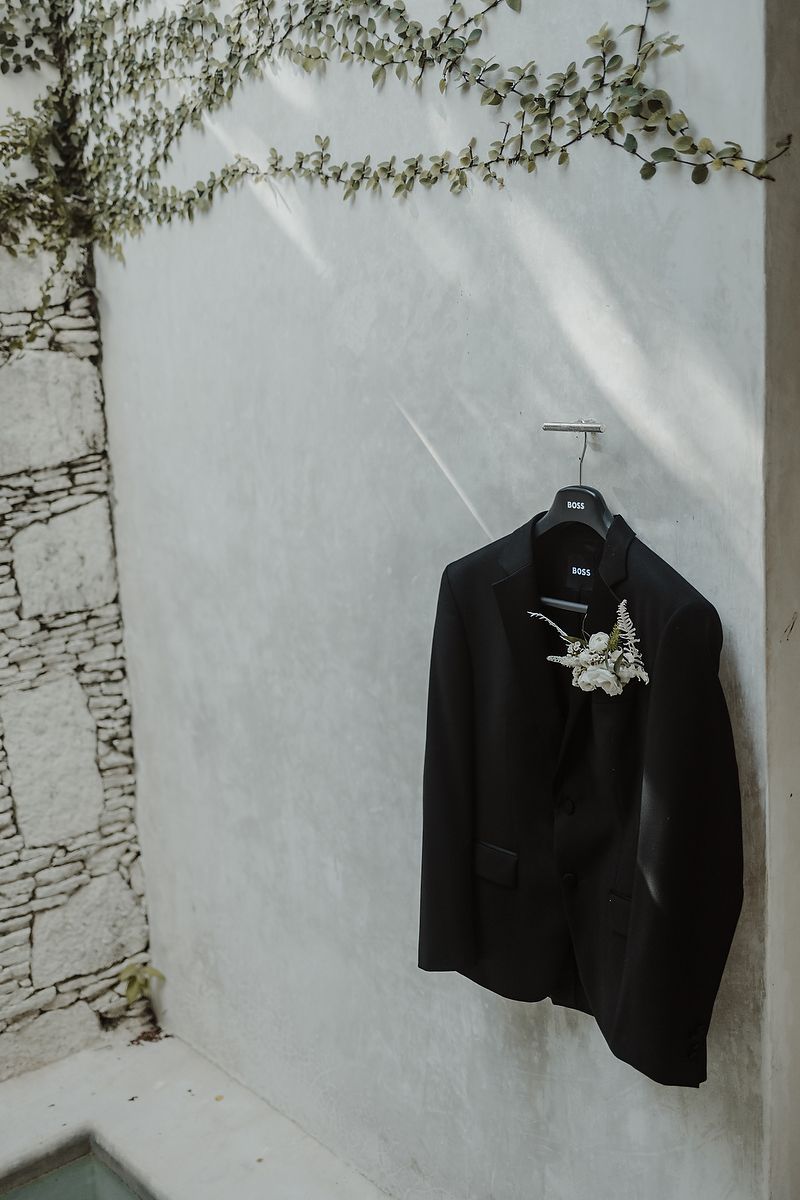 Black BOSS tuxedo jacket with white floral boutonniere on hanger against white wall with trailing greenery.